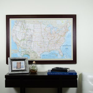 May include: A framed map of the United States of America hangs on a wall above a dark brown wooden table. The map is in a brown frame and shows the states and major cities. On the table are a silver picture frame with a photo of the Leaning Tower of Pisa, a glass vase with a plant, a black notebook, and a blue book titled "World Reference Atlas".