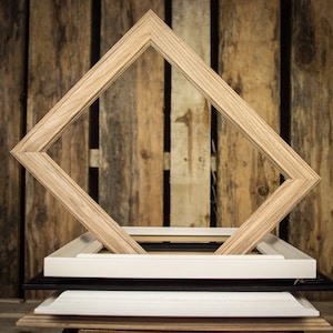 May include: A stack of four picture frames, each with a different color and style. The top frame is a light wood diamond shape, the second is white, the third is black, and the bottom is a light wood.