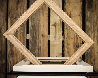 May include: A stack of four picture frames, each with a different color and style. The top frame is a light wood diamond shape, the second is white, the third is black, and the bottom is a light wood.