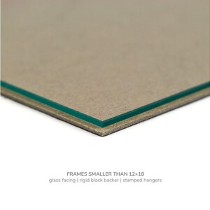 May include: Close-up of the edge of a picture frame showing a glass facing, a rigid black backer, and stamped hangers. FRAMES SMALLER THAN 12x18.