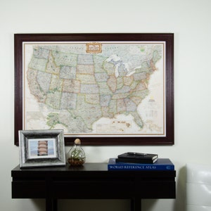 May include: A framed map of the United States with a brown frame. The map is detailed and shows state borders, major cities, and geographical features. The map is hanging on a white wall above a dark brown table with a silver picture frame, a clear glass vase with a plant, a black notebook, and a blue book titled "World Reference Atlas".