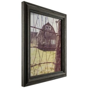 May include: A framed photograph of a rustic barn seen through a wire fence. The barn is painted a dark red with white windows and is set against a backdrop of a green field and a blue sky. The photograph is framed in a dark gray wood frame.