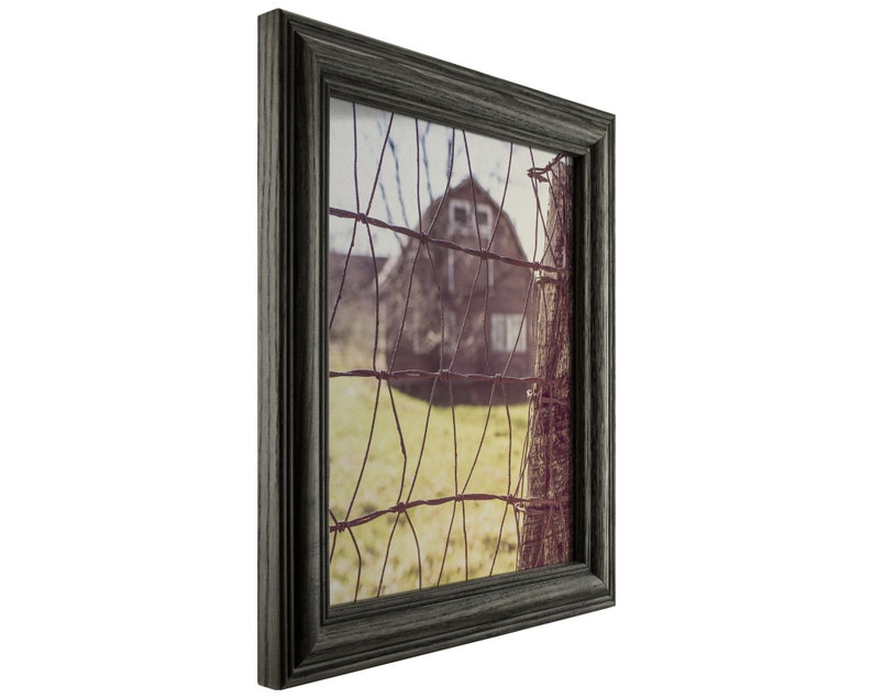 May include: A framed photograph of a rustic barn seen through a wire fence. The barn is painted a dark red with white windows and is set against a backdrop of a green field and a blue sky. The photograph is framed in a dark gray wood frame.