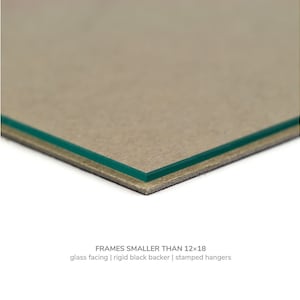 May include: Close-up of the edge of a picture frame showing a glass facing, a rigid black backer, and stamped hangers. FRAMES SMALLER THAN 12x18.
