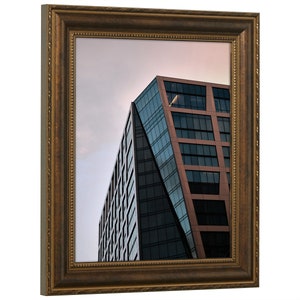 Pu&ograve; includere: Una fotografia incorniciata di un edificio moderno con una facciata in vetro e metallo. L'edificio &egrave; inclinato e ha una superficie riflettente, che riflette il cielo e le nuvole. La cornice &egrave; in legno marrone scuro con un bordo decorativo.