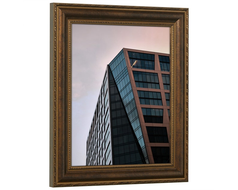 Pu&ograve; includere: Una fotografia incorniciata di un edificio moderno con una facciata in vetro e metallo. L'edificio &egrave; inclinato e ha una superficie riflettente, che riflette il cielo e le nuvole. La cornice &egrave; in legno marrone scuro con un bordo decorativo.