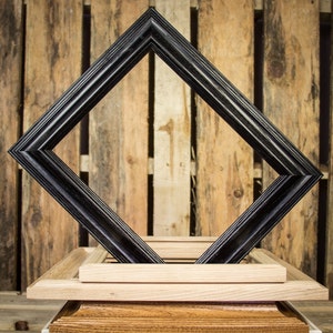 May include: A black wood picture frame with a beveled edge, sitting on top of a stack of three wooden frames. The frames are all different sizes and are made of different types of wood.