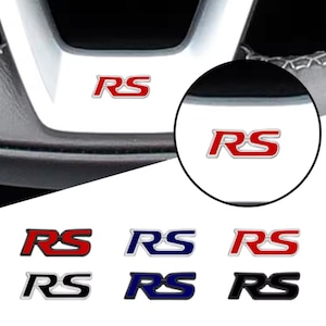 May include: A collection of RS emblems featuring the letters R and S in a stylised font. The set includes six individual badges in various colour combinations, including red, blue, black, and silver, shown applied to a steering wheel surface.