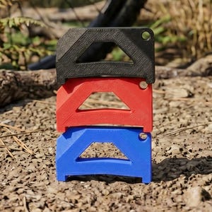 Könnte beinhalten: Drei gestapelte, farbenfrohe Fahrradrahmenschützer. Der obere Schützer ist schwarz, der mittlere rot und der untere blau. Jeder hat einen dreieckigen Ausschnitt und ein kleines Loch. Die Schützer liegen auf einem Bett aus kleinen Steinen mit verschwommenem Hintergrund.