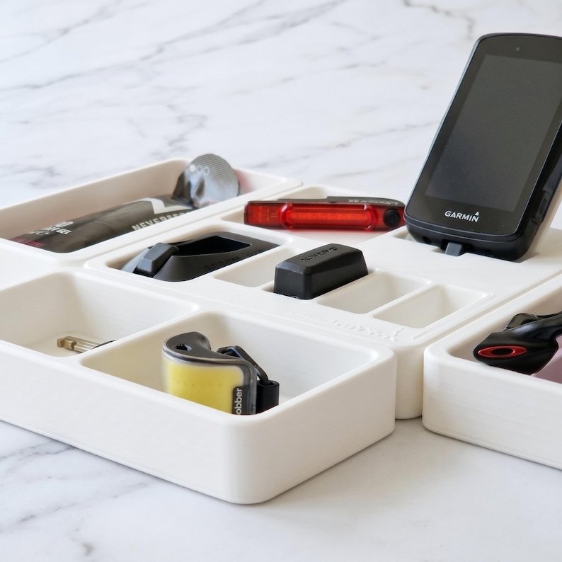 May include: A white compartmentalized organizer tray holding various small items. Items include a black Garmin device, a red light, and other black accessories. The tray is on a white marble surface.
