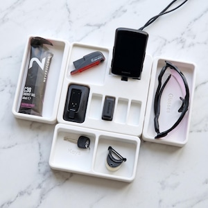 May include: A white organizer tray with various items, including a Garmin device, sunglasses, keys, and energy gel. The tray also holds a red bike light and other small black accessories. The energy gel package reads "C30 Energy Gel 40ml e Passion Fruit."