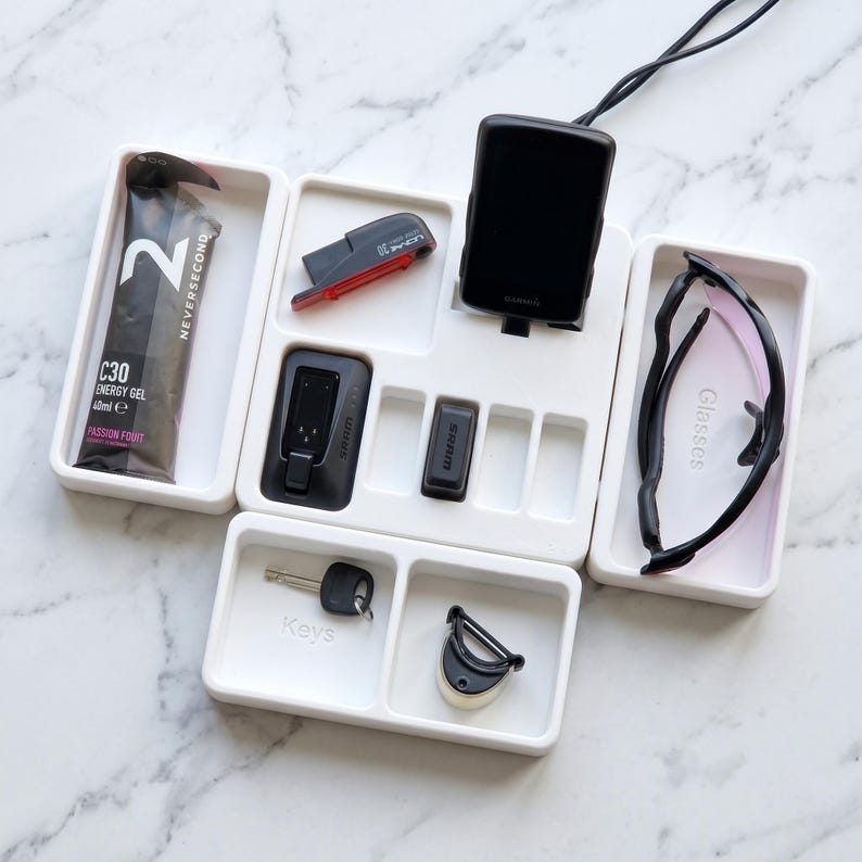 May include: A white organizer tray with various items, including a Garmin device, sunglasses, keys, and energy gel. The tray also holds a red bike light and other small black accessories. The energy gel package reads "C30 Energy Gel 40ml e Passion Fruit."