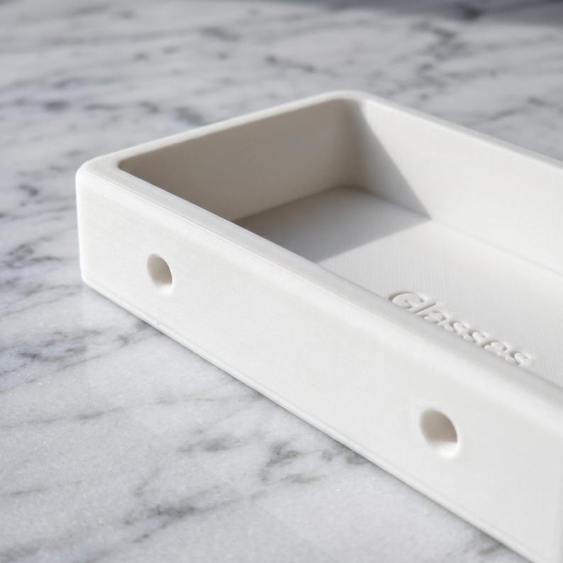 May include: A white rectangular glasses holder with rounded corners and two small holes on the side. The word "Glasses" is embossed on the inside bottom of the holder. The holder is made of a smooth, solid material.