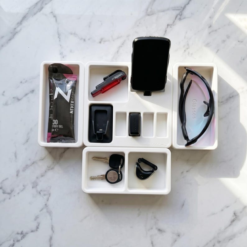 May include: A white organizer tray with compartments holding various items. Includes a black energy gel packet, a red and black bike light, a black device, sunglasses, keys, and a black clip. The tray is on a marble surface.