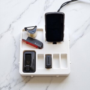 May include: A white organizer tray holding various cycling accessories. Items include a black cycling computer, a red and black rear light, a small silver light, and two black SRAM components. The tray is on a marble surface.