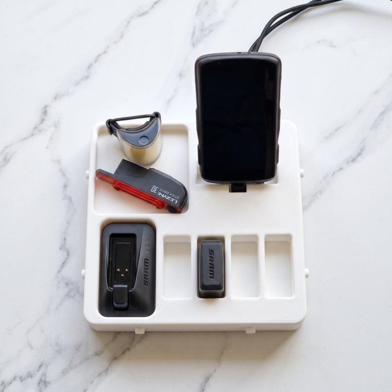 May include: A white organizer tray holding various cycling accessories. Items include a black cycling computer, a red and black rear light, a small silver light, and two black SRAM components. The tray is on a marble surface.