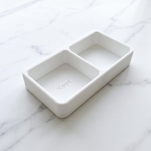 May include: A white rectangular tray with two compartments. The word "Keys" is subtly printed in the bottom of one compartment. The tray is made of a smooth, matte material and is designed for organizing small items.