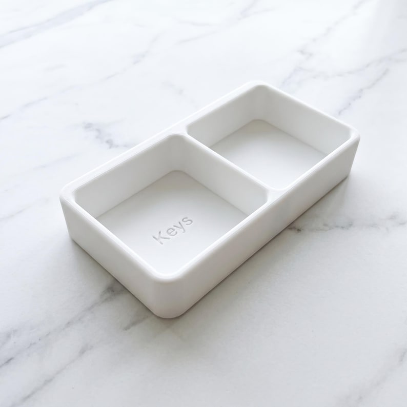 May include: A white rectangular tray with two compartments. The word "Keys" is subtly printed in the bottom of one compartment. The tray is made of a smooth, matte material and is designed for organizing small items.
