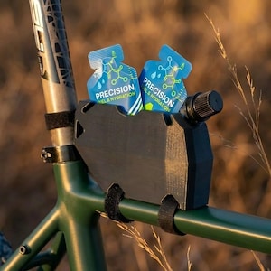K&ouml;nnte beinhalten: Ein schwarzes Fahrradzubeh&ouml;r h&auml;lt zwei blau-wei&szlig;e P&auml;ckchen mit der Aufschrift "Precision Fuel & Hydration". Das Zubeh&ouml;r ist an einem gr&uuml;nen Fahrradrahmen mit schwarzen Klettb&auml;ndern befestigt. Der Hintergrund ist verschwommen mit trockenem Gras.