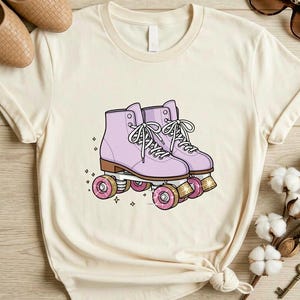 May include: Cream-colored t-shirt featuring a graphic of lavender roller skates with pink wheels. The skates have white laces and are set against a backdrop of small stars. The word "NATURAL" is printed at the bottom left.