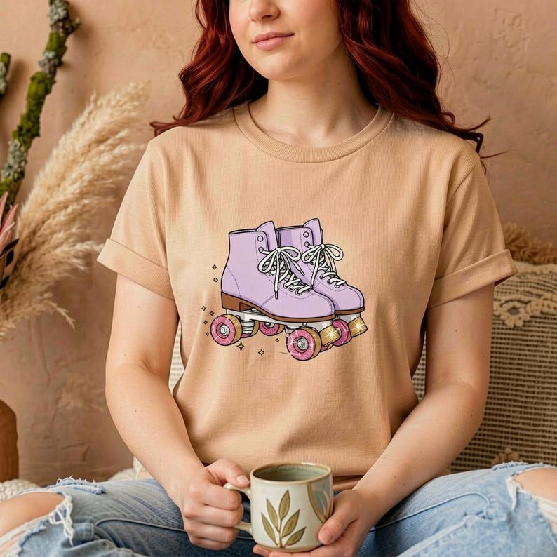 May include: A tan t-shirt featuring a graphic of a pair of lavender roller skates with pink wheels. The skates have white laces and are set against a neutral background. The shirt is a casual, everyday wear item.