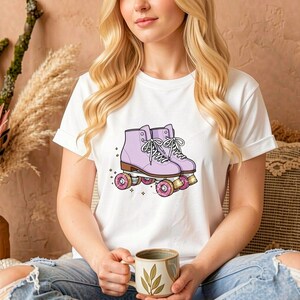 May include: White t-shirt featuring a graphic of lavender roller skates with pink wheels and white laces. The skates are detailed with brown soles and are set against a backdrop of small stars. The shirt is a classic crew neck.