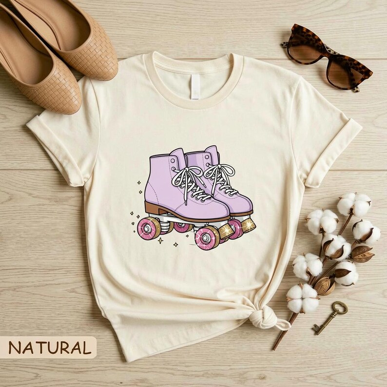 May include: Cream-colored t-shirt featuring a graphic of lavender roller skates with pink wheels. The skates have white laces and are set against a backdrop of small stars. The word "NATURAL" is printed at the bottom left.