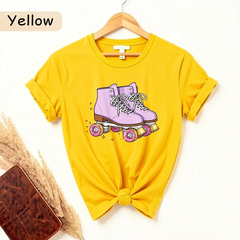 May include: Yellow t-shirt featuring a graphic of pink roller skates with white laces and pink wheels. The word "Yellow" is in the top left corner. The shirt is knotted at the waist and hangs on a wooden hanger.