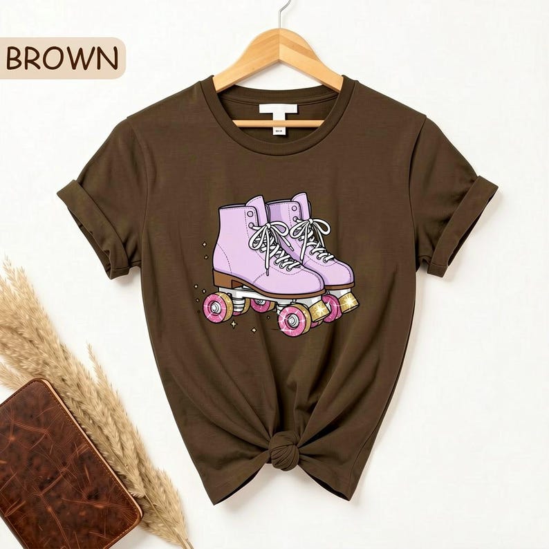 May include: Brown t-shirt featuring a graphic of pink roller skates with white laces and pink wheels. The word "BROWN" is in a brown box in the upper left corner. The shirt is hanging on a wooden hanger.