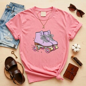 May include: A pink t-shirt featuring a graphic of lavender roller skates with white laces and pink wheels. The shirt is styled with denim shorts, brown sandals, and a gold necklace. Other accessories include sunglasses and a small leather-bound journal.