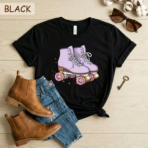 May include: Black t-shirt featuring a graphic of lavender roller skates with pink donut wheels. The shirt is paired with blue jeans and brown ankle boots. The word "BLACK" is printed at the top left.
