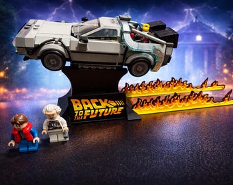 May include: A grey plastic building block model of a sports car mounted on a black display stand with the text BACK TO THE FUTURE. Two small plastic figures stand in front, accompanied by two translucent orange flame pieces on the ground.
