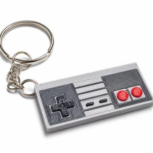 May include: A rectangular keyring shaped like a retro video game controller. The device is light grey with a dark grey directional pad, two small black buttons, and two circular red buttons on the right side, attached to a silver metal ring.