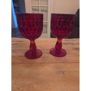 Puede incluir: Dos copas de vino rojo rubí con un patrón texturizado. Las copas tienen una base redonda, un tallo y una copa. Las copas son de vidrio y están sobre una superficie de madera.