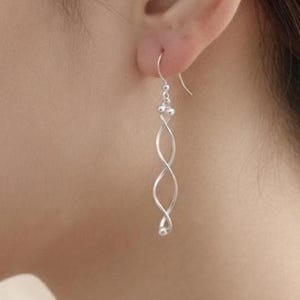 Könnte beinhalten: Ein silberner Hängeohrring mit einem zarten, länglichen Spiraldesign. Der Ohrring hängt an einem einfachen Haken und verfügt über kleine kugelförmige Akzente an der Ober- und Unterseite der gedrehten Metallstruktur für eine elegante Optik.