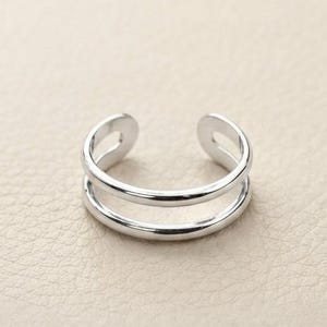 Könnte beinhalten: Ein polierter silberfarbener Ring mit offenem Design, bestehend aus zwei parallelen Bändern, die an den Enden verbunden sind. Das Metall hat eine glatte, reflektierende Oberfläche, und die Öffnung ermöglicht eine Anpassung der Ringgröße.