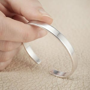 Könnte beinhalten: Ein glatter, polierter silberfarbener Armreif mit einem einfachen, offenen Design. Das Metallband weist eine saubere, reflektierende Oberfläche und eine gleichmäßige Breite auf und bildet eine kreisförmige Kurve für das Handgelenk.