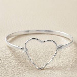 May include: A silver-toned bangle bracelet featuring a central open heart-shaped design. The bracelet has a smooth, polished finish and includes a small, coiled wire detail on one side where the heart shape connects to the circular band.
