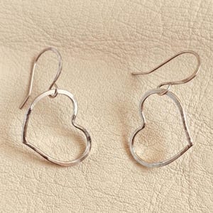 Könnte beinhalten: Ein Paar silberfarbene Metallohrringe mit offenem herzförmigem Design. Jeder Ohrring hängt an einem geschwungenen Haken und zeigt eine einfache, dünne und polierte metallische Kontur in Form eines Herzens mit einer glatten Oberfläche.