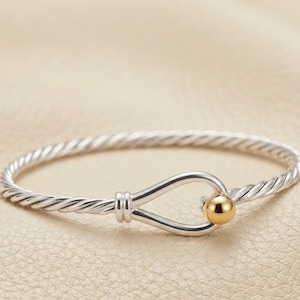 Könnte beinhalten: Ein silberfarbenes Armband mit gedrehtem Kabeldesign. Das Armband verfügt über einen Verschluss im Haken-Stil, der in einer glatten, polierten goldfarbenen Kugel endet und so einen sicheren und markanten Blickfang bildet.