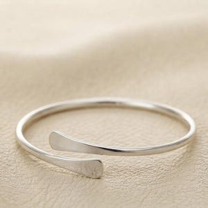 Könnte beinhalten: Ein silberfarbenes, offenes Armband mit einem dünnen, kreisförmigen Ring, der in zwei flache, abgerundete Enden ausläuft, die sich nicht berühren. Das Metall hat eine glatte, polierte Oberfläche, die entlang des gesamten Stücks reflektiert.