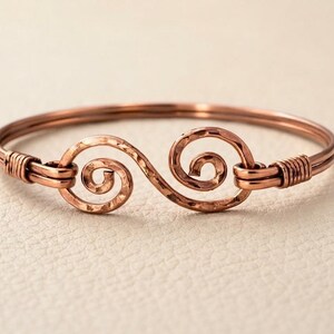 Puede incluir: Una pulsera de alambre de cobre con un diseño central de doble espiral. La superficie del metal tiene una textura martillada y los extremos del alambre están envueltos firmemente alrededor de la banda principal para asegurar el punto focal.