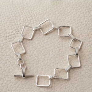 Könnte beinhalten: Ein silberfarbenes Armband, das aus einer Reihe von miteinander verbundenen quadratischen Gliedern besteht. Das Armband verfügt über einen Knebelverschluss, der aus einem runden Ring und einem geraden Stab besteht.