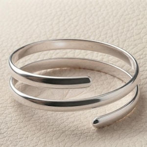 Könnte beinhalten: Ein polierter silberfarbener Bypass-Ring mit einem glatten, durchgehenden Band, das sich in einem Spiraldesign zweimal umwickelt. Das Metall hat eine reflektierende, hochglänzende Oberfläche mit abgerundeten Enden und eine klare Form.