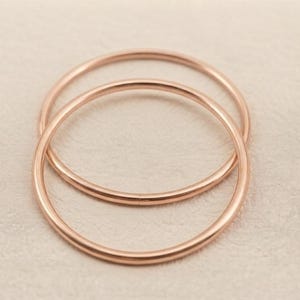 Könnte beinhalten: Zwei dünne, kreisförmige Ringe aus Roségold-Metall. Die Ringe haben eine glatte, polierte Oberfläche mit einem abgerundeten Profil. Beide Bänder sind im Design und in der Farbe identisch und zeigen einen warmen, metallischen Roségoldton.