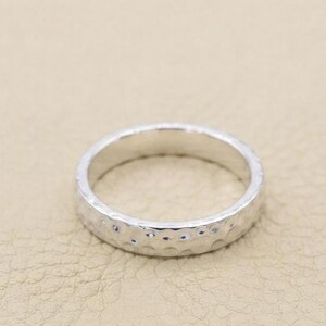 Könnte beinhalten: Ein silberfarbener Ring mit einer gehämmerten Textur auf der Außenfläche. Das Band hat eine gleichmäßige Breite und eine reflektierende metallische Oberfläche, die zahlreiche kleine, kreisförmige Vertiefungen aufweist.