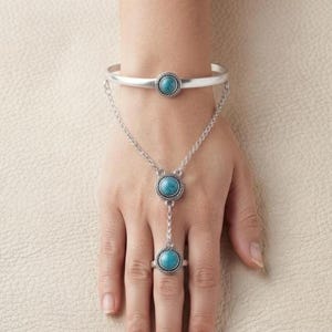 Könnte beinhalten: Ein silberfarbenes Handkettchen mit einem kreisförmigen, türkisfarbenen Stein in einer seilartig texturierten Fassung am Armband, das durch eine feine Kette mit einem passenden Stein am Fingerring verbunden ist und ein stimmiges Schmuckstück bildet.