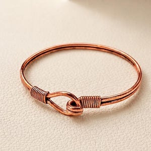 Könnte beinhalten: Ein kreisförmiges Armband aus Kupfer mit einem Haken- und Ösenverschluss. Das Metall hat ein warmes, rotbraunes metallisches Finish mit sichtbaren, drahtumwickelten Details an den Stellen, an denen der Haken in die Öse greift, was ein schlichtes Design ergibt.