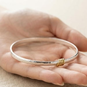 May include: A silver bangle bracelet with a hammered texture and a polished finish. The circular band features a small, gold-colored accent consisting of three thin, coiled wire rings wrapped around the silver surface of the bangle.