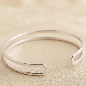 Könnte beinhalten: Ein silberfarbenes Armband in Manschettenform mit einem Doppelband-Design und einer gehämmerten, strukturierten Oberfläche. Der offene Armreif besteht aus zwei parallelen Metallstreifen, die sich zu einer abgerundeten Form biegen.